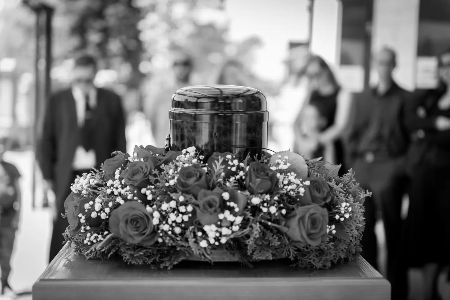 Urne im Blumenkranz bei Begräbnis mit Trauernden im Hingergrund, begleitet von Trauerrednerin