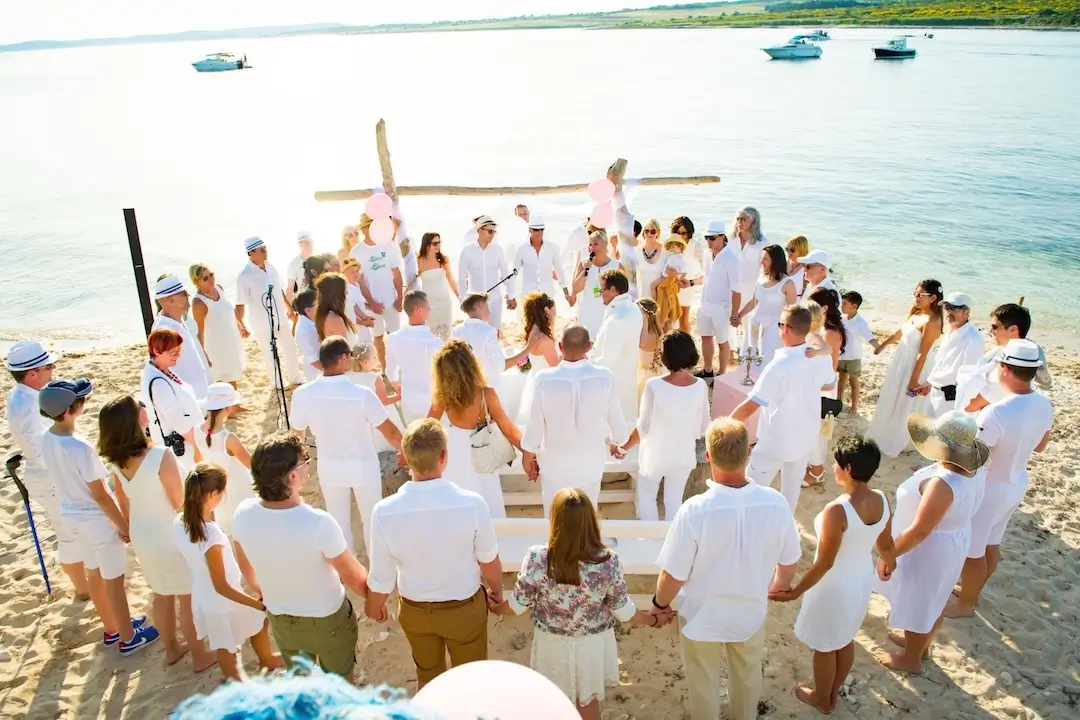 Gäste in Weiß beim Jawort am Meer stehen im Kreis und halten sich an den Händen
