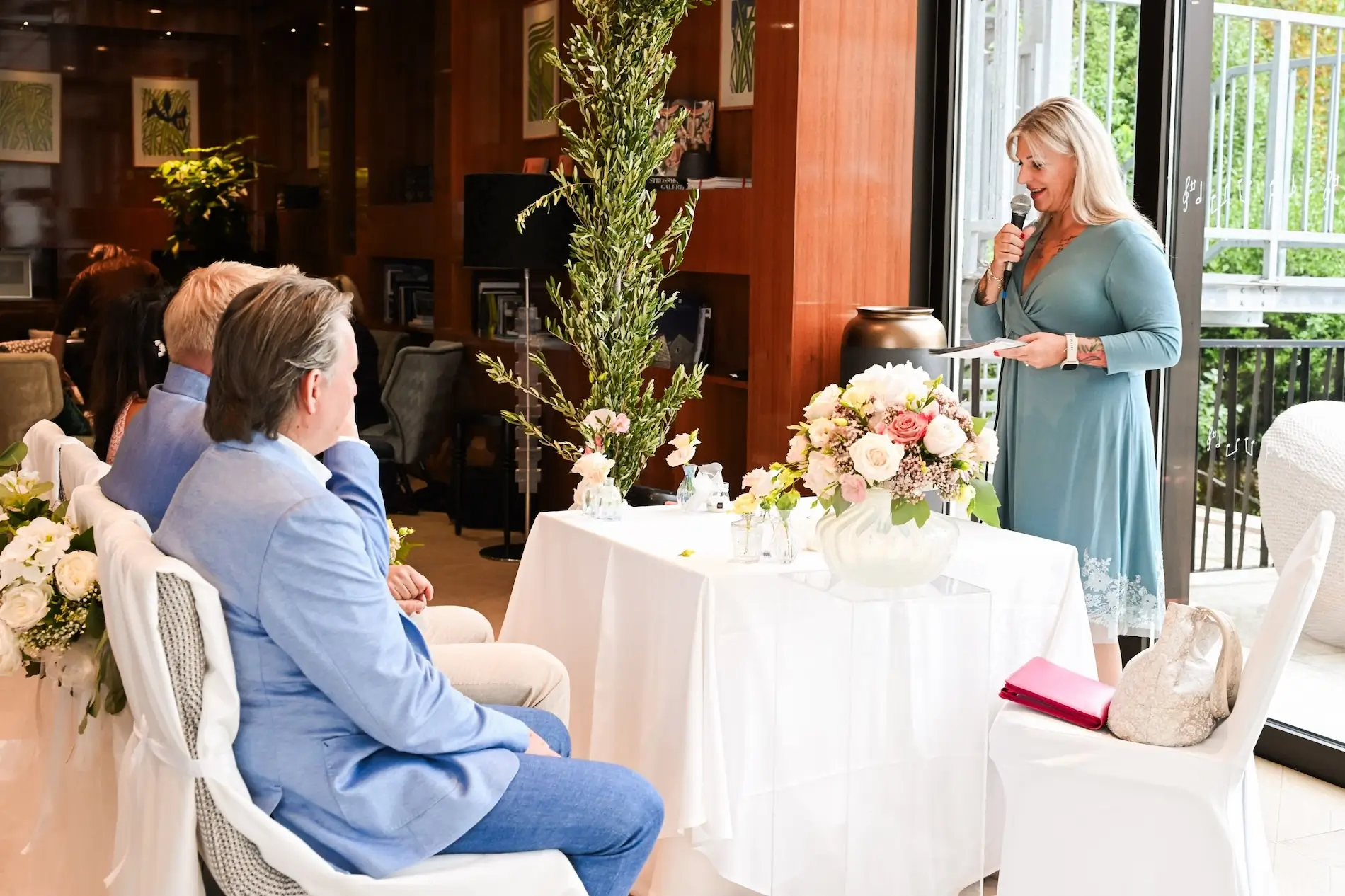 Heike Schönbacher in hellem Raum mit Handmikrofon hinter Tisch und Blumenstrauß bei der freien Trauung