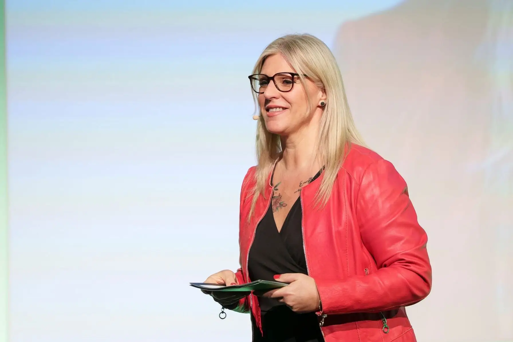 Heike Schönbacher in roter Jacke bei Moderation auf der Bühne vor großer Leinwand